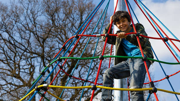 Boy smiles as he sits in a Hullabazoo Adventure Play climbing frame 