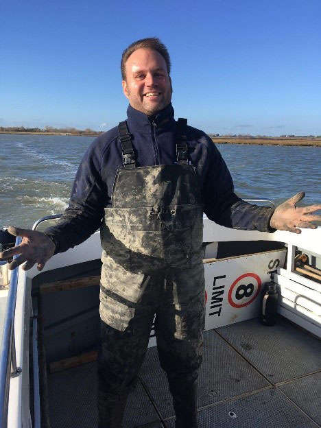 Photograph of Ken on the boat, wearing waders and covered in mud.
