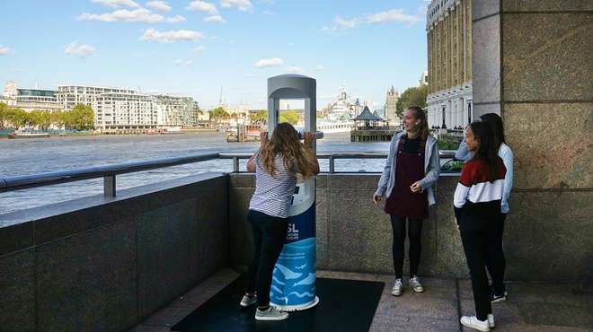 Photo - A girl trying the VR experience while her friends watch, with the Thames and Tower Bridge in the background