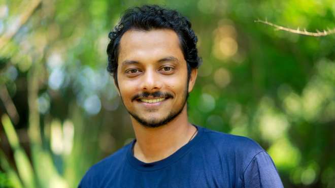 Photo - Portrait shot of Rajkumar smiling at the camera, wearing a blue t-shirt with greenery in the background.