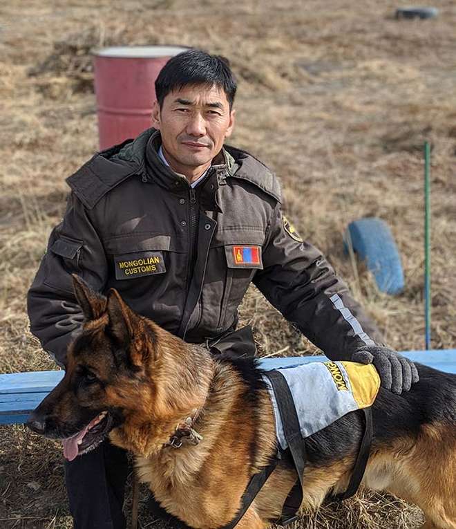 Photo - A Mongolian Customs Officer in uniform kneeling on the grass outside with his detector dog