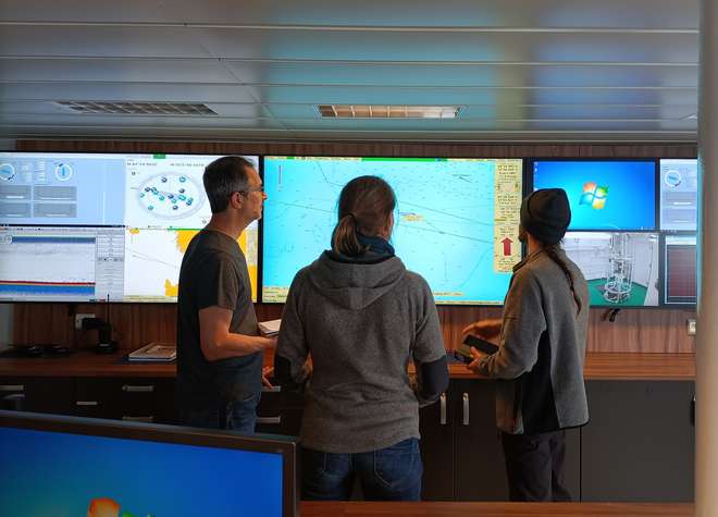 Photo of Julian, Nanette and Chris standing inside the ship, next to some large screen monitors.