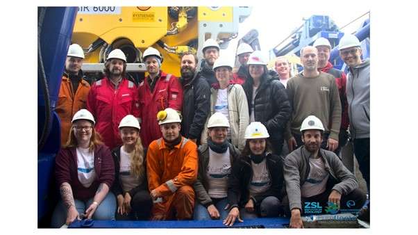 The team of scientists, ROV pilots and technicians, all wearing hard hats onboard the research vessel, smile for the camera.